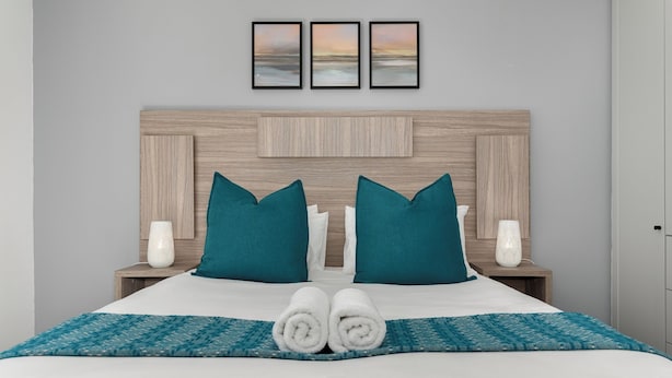 A neatly made bed with white linens, teal pillows, and a teal-patterned throw. There are two white vases on bedside tables, and three framed pictures above the bed.