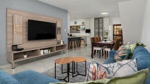 A modern living room with a flat-screen TV, wooden entertainment unit, blue sofa, and a dining area with a wooden table and chairs.