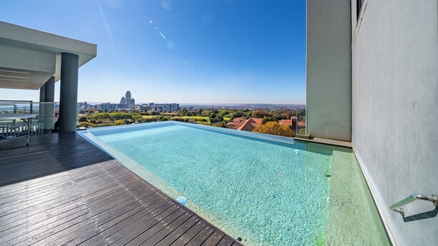 Une piscine extérieure moderne, dotée d’une terrasse en bois, offrant une vue dégagée sur le paysage urbain et laissant apercevoir un grand bâtiment au loin.