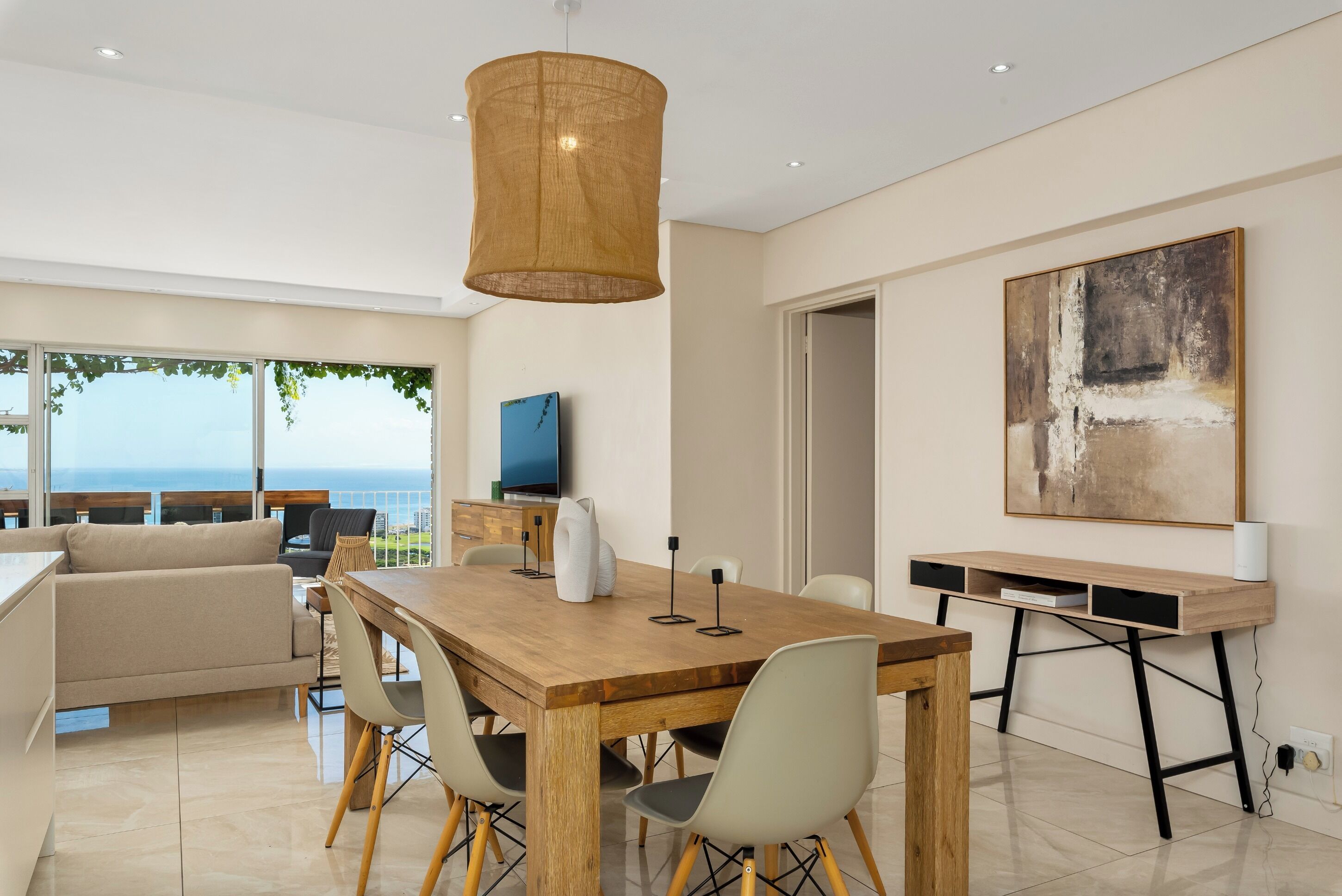 A modern living room with a wooden dining table, beige sofa, and a large window overlooking the sea.