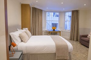 A hotel room with a large bed, a small table, a sofa, and a window with curtains. - The Courtfield by Nestor - Studio LG Kensington (London)