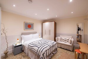 A bedroom with a bed, nightstand, bedside lamp, wardrobe, sofa, and a small table. - The Courtfield - Studio Lower Ground in Kensington (London)