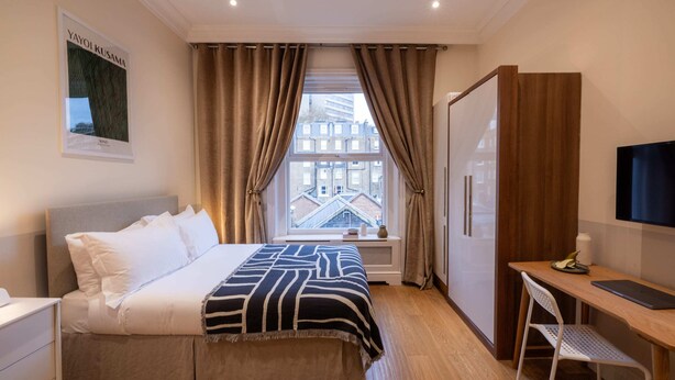 A hotel room with a bed, a desk, a chair, a TV, and a window with curtains. - The Courtfield - Studio Apartment in Kensington (London)