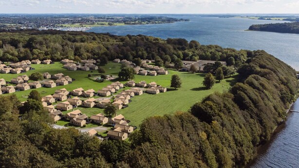 Luchtbeeld van een kustdorp, met huizen in een halve cirkel langs de kustlijn gelegen, omringd door weelderige begroeiing en een groot wateroppervlak.