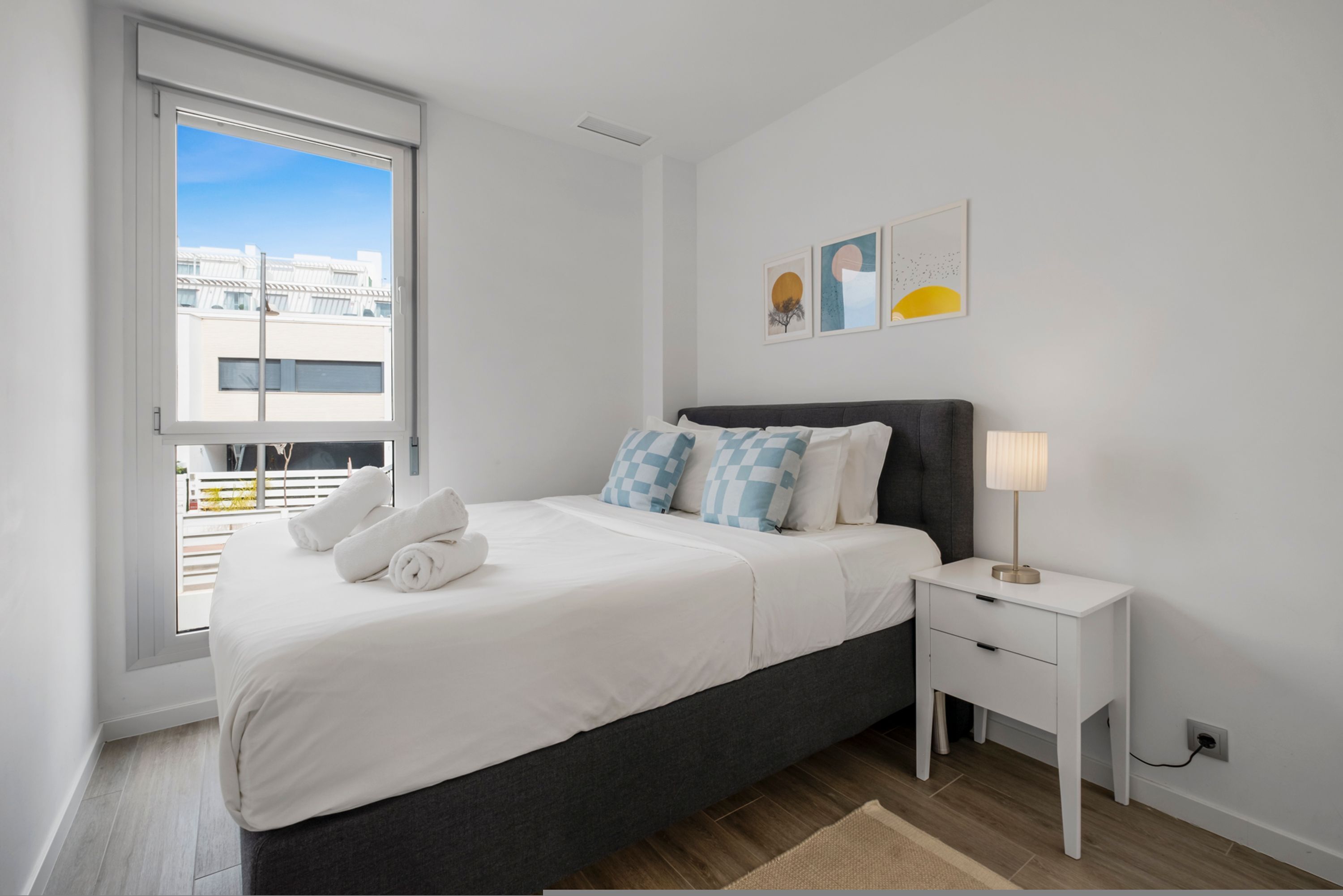 A modern bedroom with a large bed, a bedside table, and a window with a view of buildings.