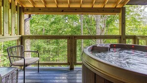 Una terrazza in legno con vasca idromassaggio e una sedia in vimini, con vista su una zona boschiva.