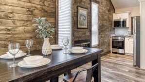 Zona pranzo con tavolo in legno apparecchiata per quattro persone, finestre con persiane e cucina dotata di elettrodomestici moderni, più un quadro appeso alla parete.