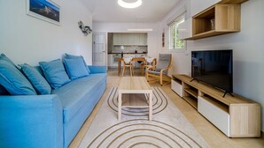 Ein modernes Wohnzimmer mit einem blauen Sofa, einem Holztisch, einem Fernseher und einer Küche im Hintergrund.
