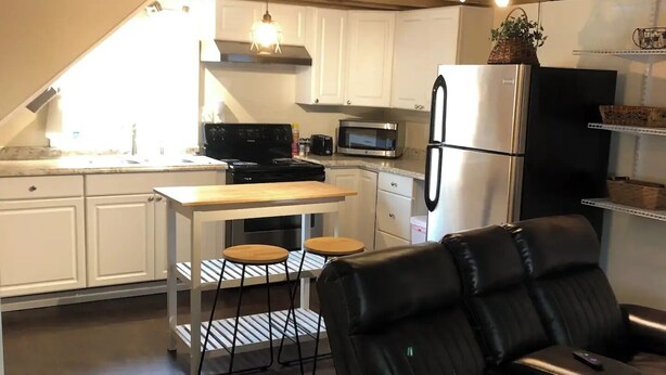 Et moderne køkken med hvide skabe, et sort køleskab, en mikrobølgeovn og en komfur. Der er et spisebord med to barstole og en sort lædersofa i opholdsområdet.