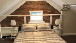 Et soveværelse med skråvæg, en murstensvæg, en seng med stribet sengetøj, to sengeborde med lamper og et vindue med udsigt til himlen.