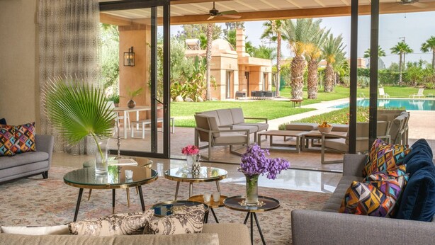 A spacious living room with a large glass door leading to a pool area, furnished with sofas, tables, and decorative plants.