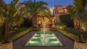 Un espace extérieur bien éclairé autour d’une piscine, agrémenté d’une fontaine, entouré d’une végétation luxuriante et bordé d’un bâtiment doté d’un balcon.