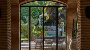 Une vue à travers une porte vitrée donnant sur un patio aménagé avec des sièges d’extérieur et un pot de fleurs.