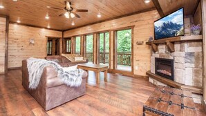 A wooden cabin interior with a sofa, a fireplace, a television, and large windows. - Epic Stay: Private Pool, Theater & Games for 30 (Sevierville)