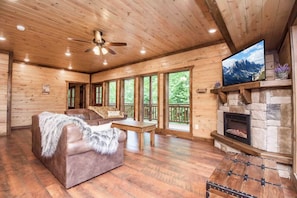 A wooden cabin interior with a sofa, a fireplace, a television, and large windows.