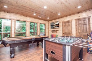 A wooden-floored room with a pool table, foosball table, and large windows.