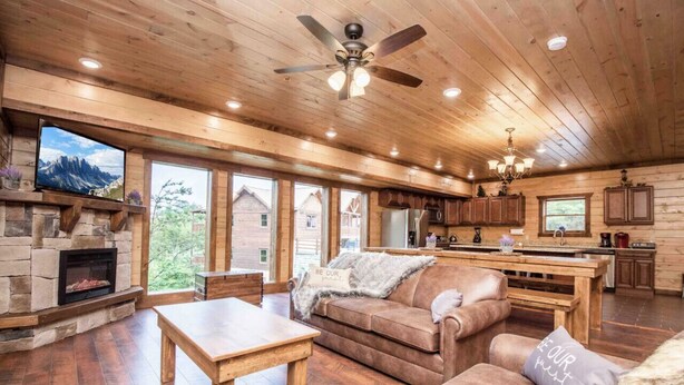 A spacious living room with a fireplace, wooden floors, a ceiling fan, and a kitchen area with wooden cabinets and a large window. - Epic Stay: Private Pool, Theater & Games for 30 (Sevierville)