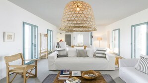 Un salon moderne avec un canapé blanc, une table basse en bois sur laquelle sont posés des livres, un luminaire suspendu et une vue sur la cuisine par une porte ouverte.