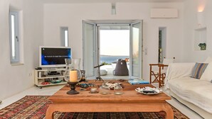 A living room with a large TV, a wooden coffee table, a sofa, and a view of the sea through a glass door. - Villa Irina - Five Bedroom Villa, Sleeps 10 (Makria Miti)