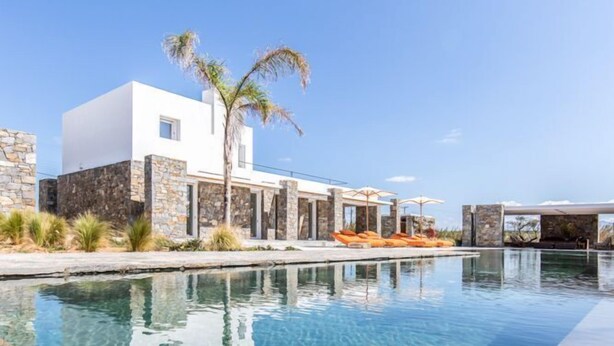 Un área de piscina moderna con agua cristalina, un edificio blanco, muros de piedra y una palmera.