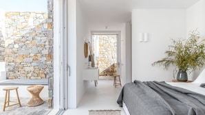 Un dormitorio moderno con una pared de piedra, una cama con ropa de cama gris, una planta en la mesita de noche y vistas a través de una puerta de cristal.