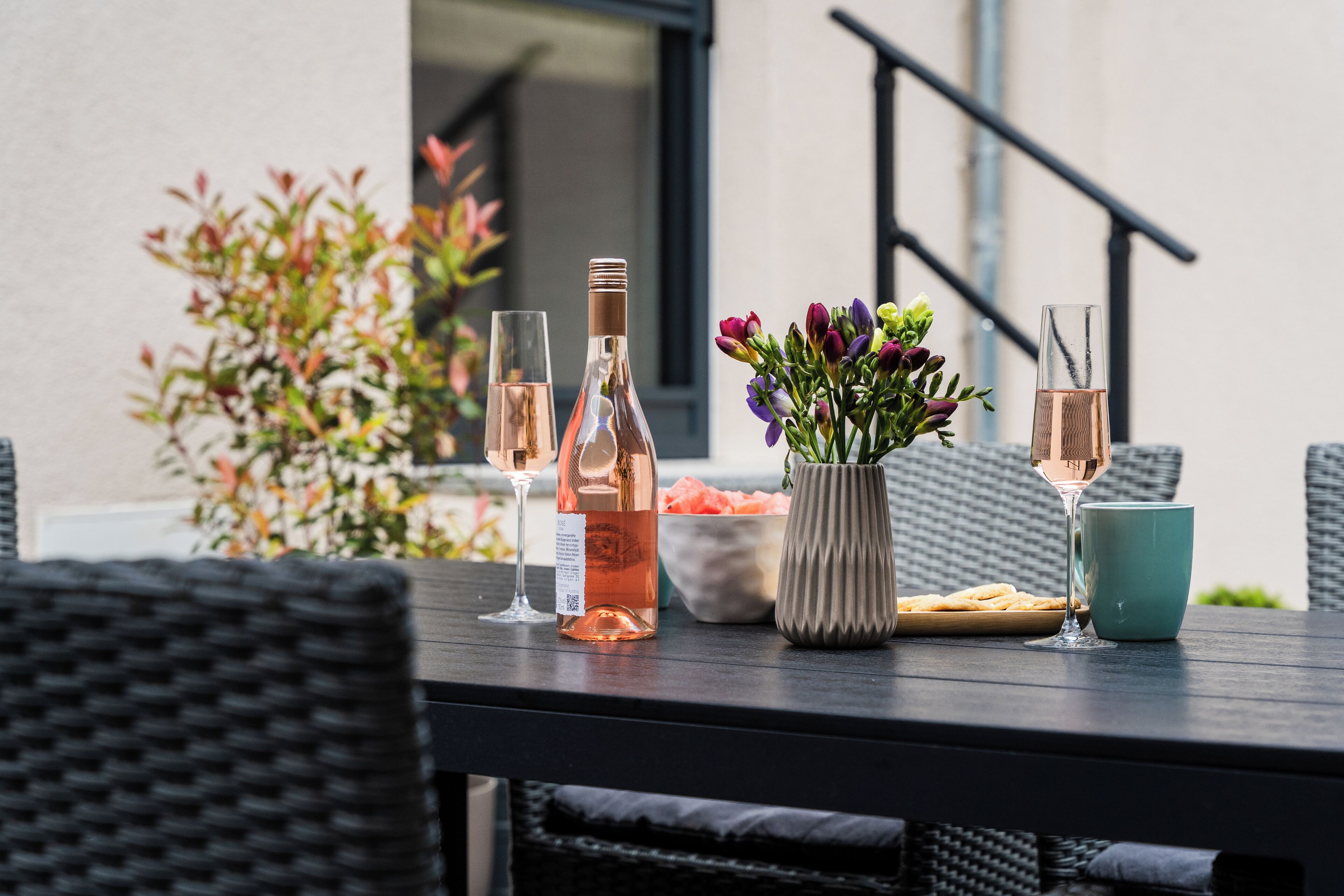 A table with two bottles of rosé wine, a glass, a vase with flowers, and a plate of food.