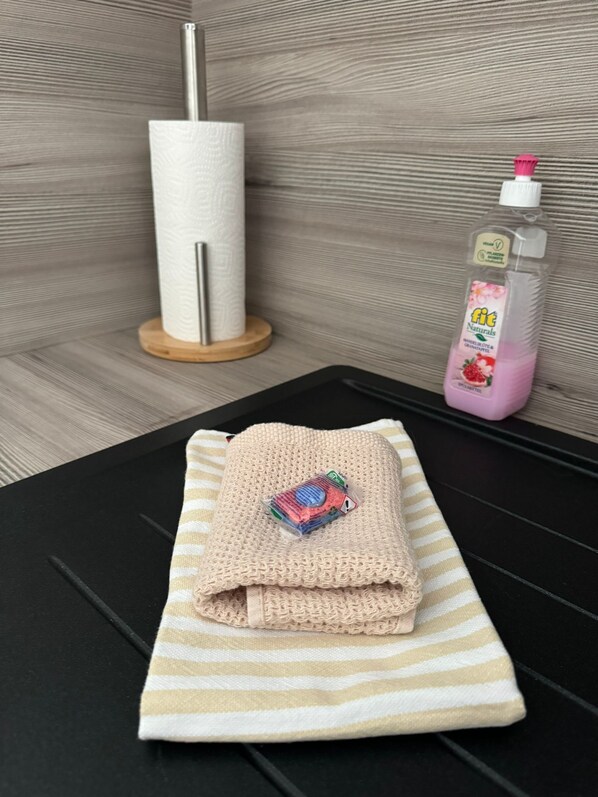 Una toalla doblada con un detalle colorido sobre un felpudo a rayas, un dispensador de toallas de papel y una botella de champú sobre una superficie negra.
