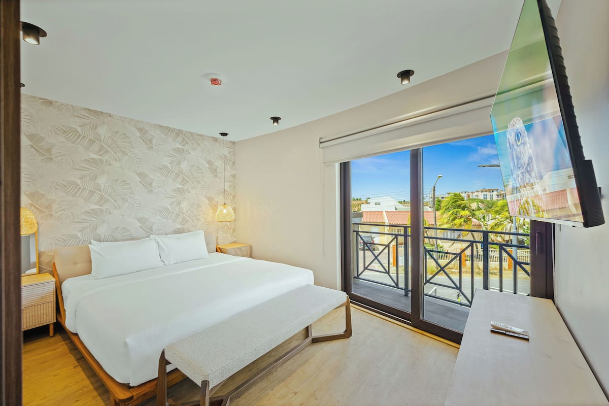 Habitación de hotel moderna con una cama grande, un escritorio y un balcón con vistas a edificios y árboles.