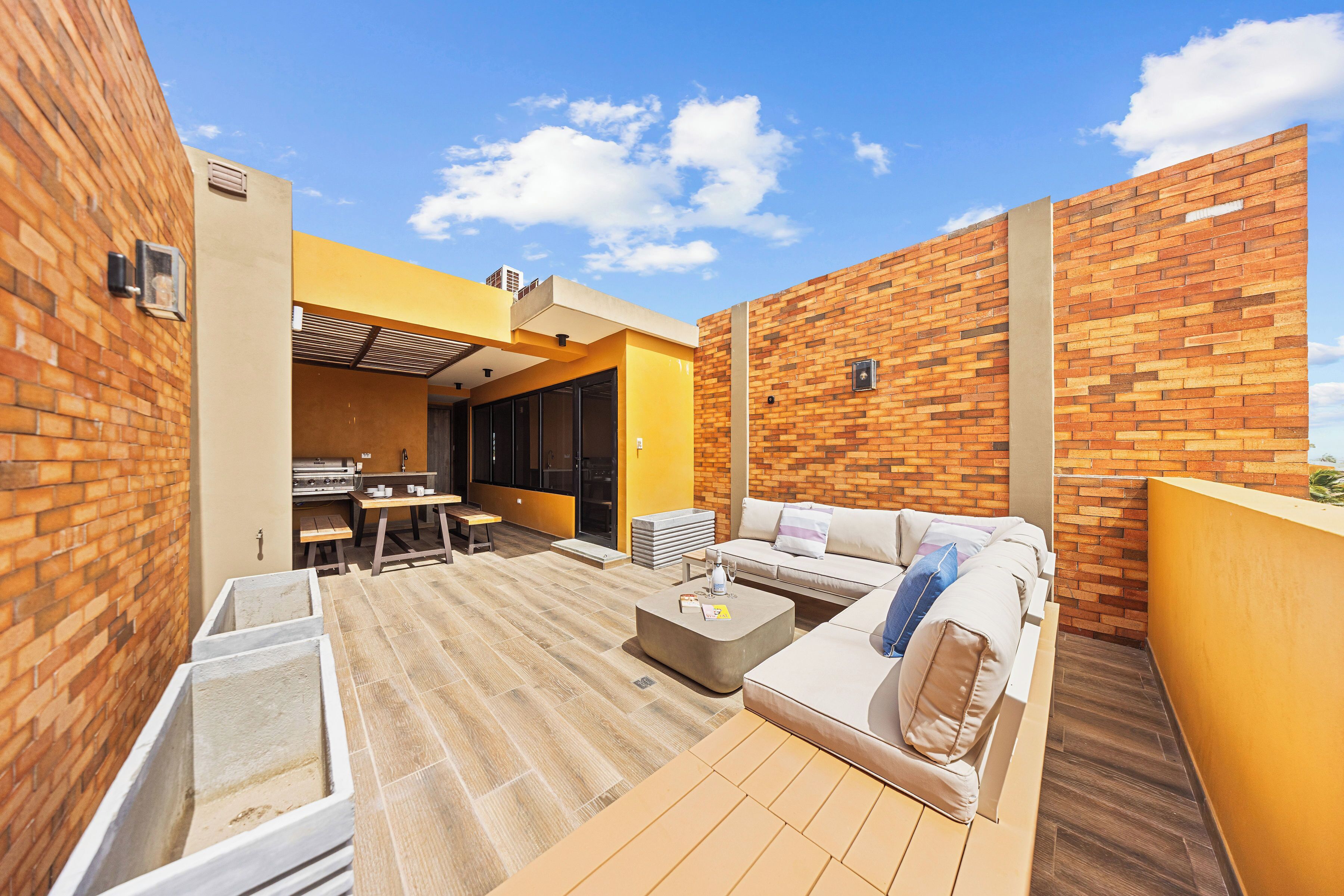 Eine Dachterrasse mit Backsteinwand, gelber Wand, Holzdeck, einer Couch und einem Grillbereich.