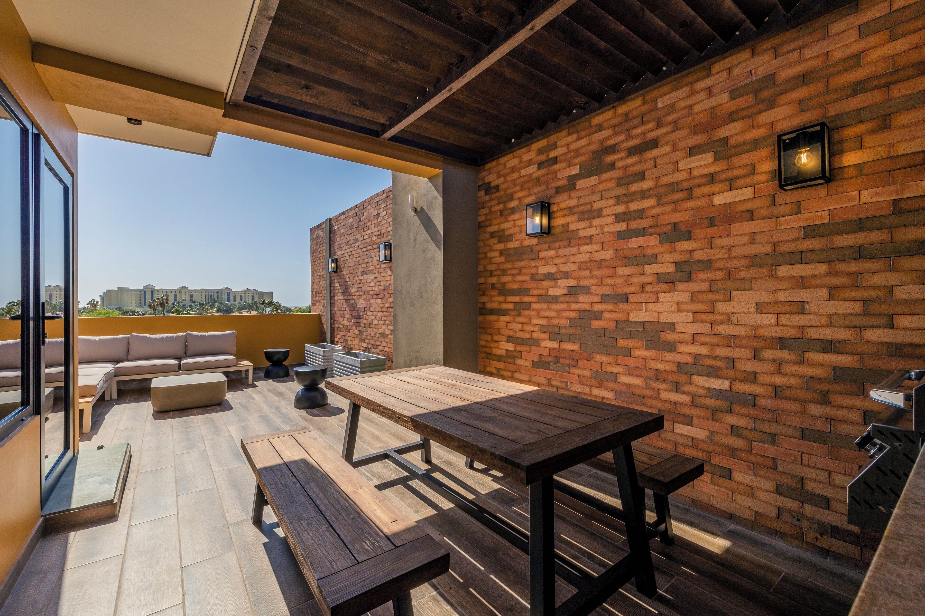An outdoor patio with a wooden table and benches, a brick wall, and a view of buildings in the distance.