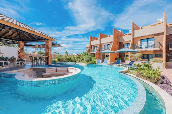 Una piscina con solárium, sillas de descanso y una zona de estar cubierta.