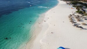 An aerial view of a beach with clear turquoise waters, white sand, and people swimming and sunbathing. - Bocobay Porta al Sole Condos (Noord)