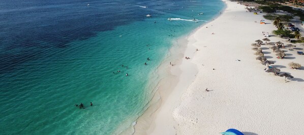 Vista aérea de una playa con aguas turquesas cristalinas, arena blanca y gente nadando y tomando el sol.