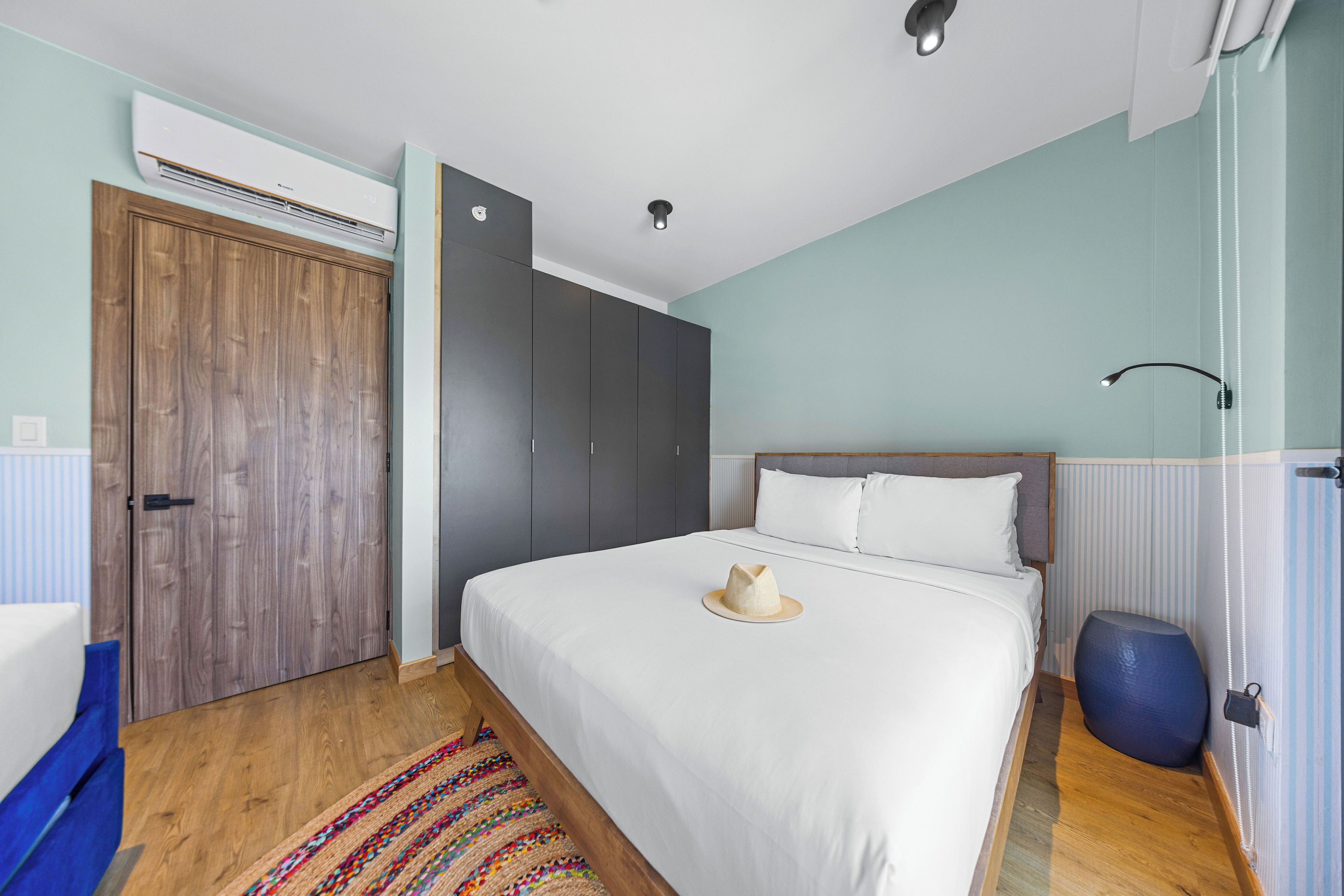 Un dormitorio con un armario de madera, una cama con ropa de cama blanca, una alfombra colorida y un reposapiés azul.