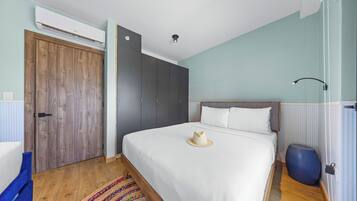 Un dormitorio con un armario de madera, una cama con ropa de cama blanca, una alfombra colorida y un reposapiés azul.