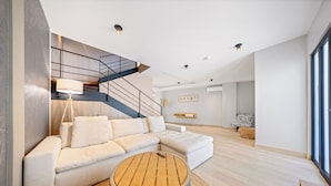 A modern living room with a sectional sofa, a round wooden coffee table, and a staircase with a black railing. - Bocobay Porta al Sole Condos (Noord)