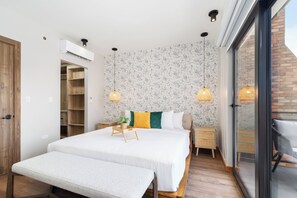 Un dormitorio con una cama grande, mesitas de noche, un banco, un equipo de aire acondicionado y una puerta corrediza de vidrio a un balcón.