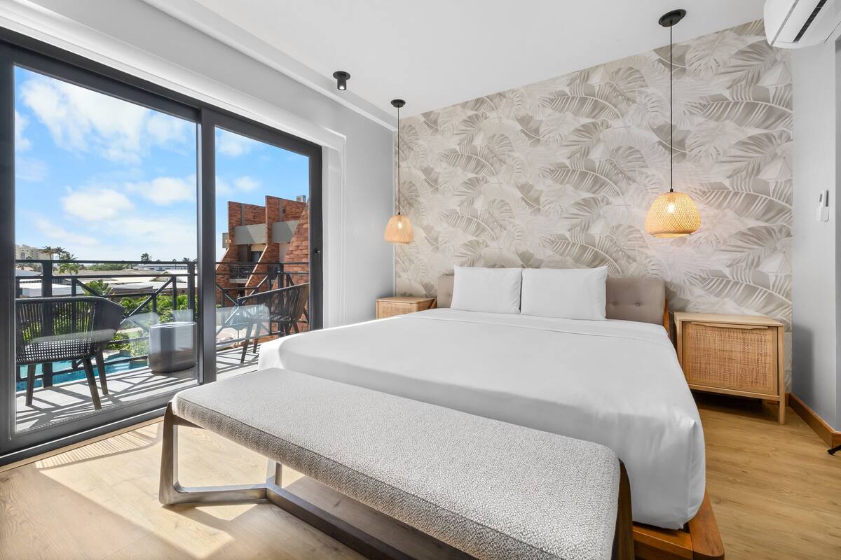 Un dormitorio moderno con una cama grande, un banco, un balcón con piscina y una pared con estampado de hojas.