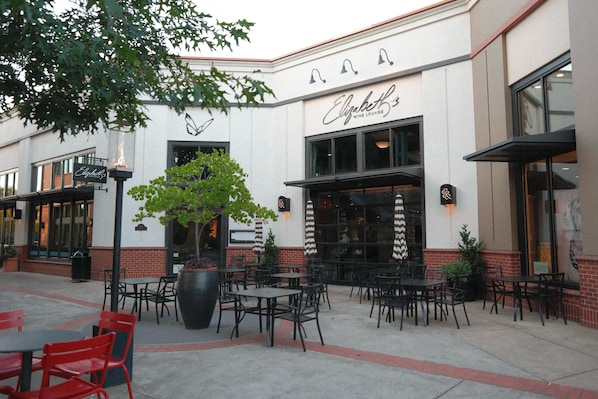 An outdoor seating area with tables and chairs, a large planter with a tree, and a building with a sign that reads 'Elizabeth's Wine Lounge'.