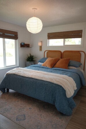 A bedroom with a bed, a hanging light fixture, a window with a view, and a bedside table with a plant.