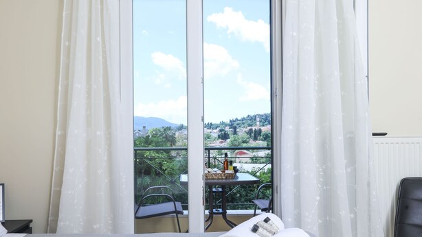 A hotel room with a bed, a table with a bottle and a cup, and a view of a balcony with chairs and a table. - Smart Home Corfu Apartment (Corfu)