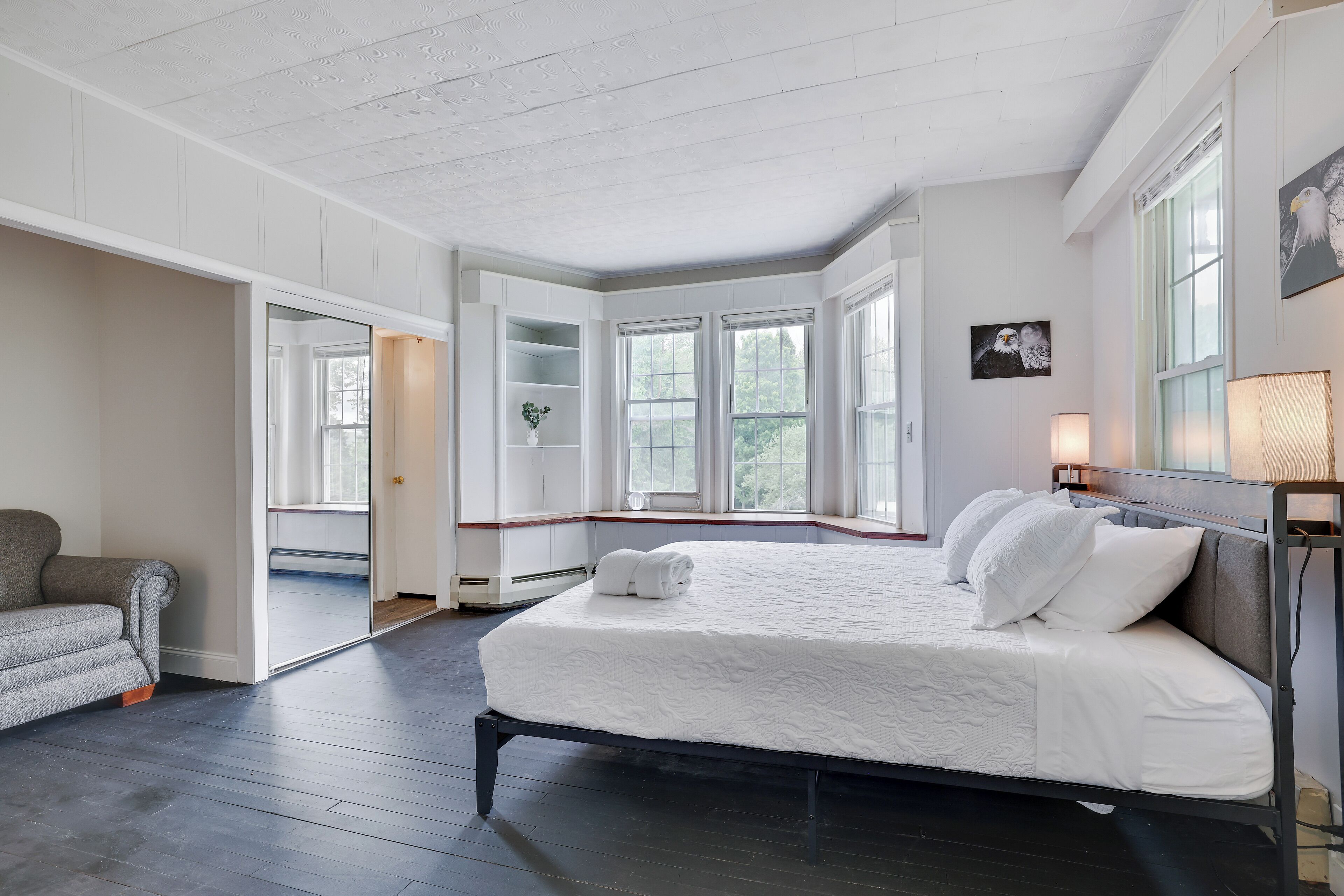 A bedroom with a large bed, a grey sofa, and a window with a view of trees.