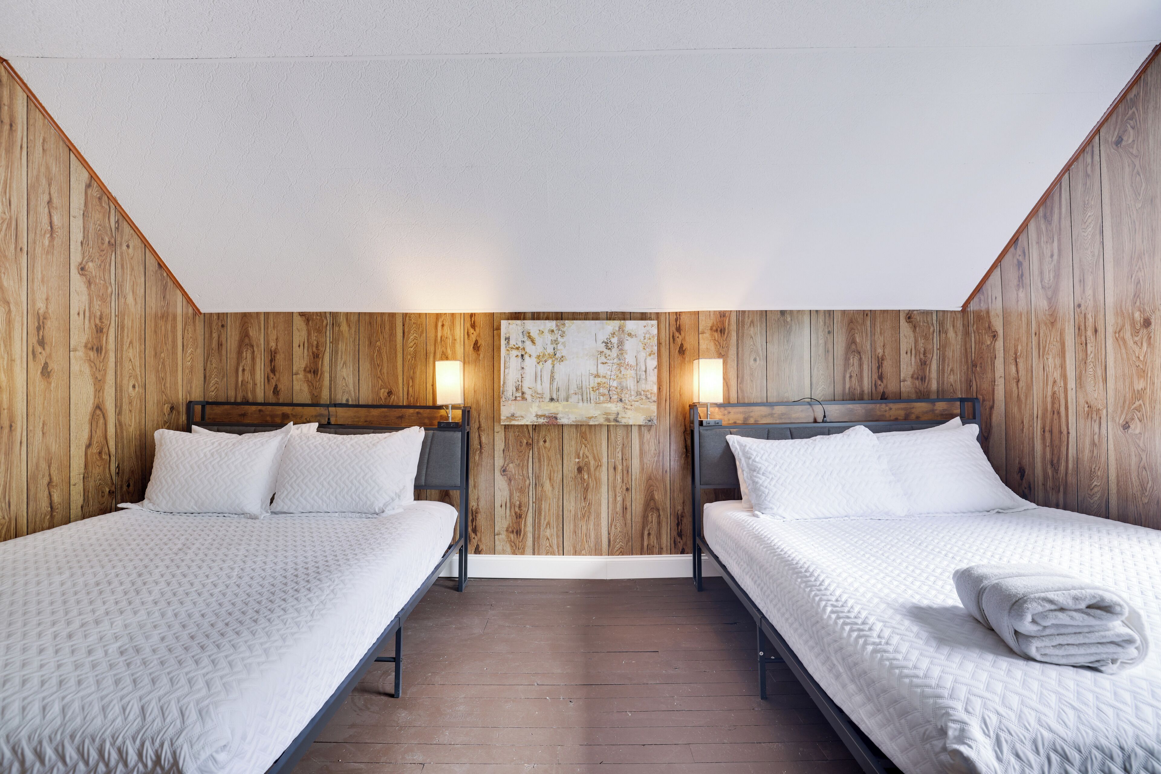 Una habitación con dos camas, paredes de madera y un cuadro en la pared trasera.