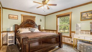 A bedroom with a wooden bed, a nightstand, a desk, and a window with a view of greenery.