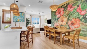 A modern kitchen with a white island, wooden dining table, and chairs. There is a large floral mural on the wall, a ceiling fan, and a window with blinds.