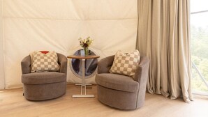 Two armchairs with checkered pillows, a round table with a vase of flowers, and a view through a window with curtains.