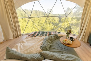 A cozy room with a large window, a bed with pillows, a blanket, and a table with a cup and a plate of food.