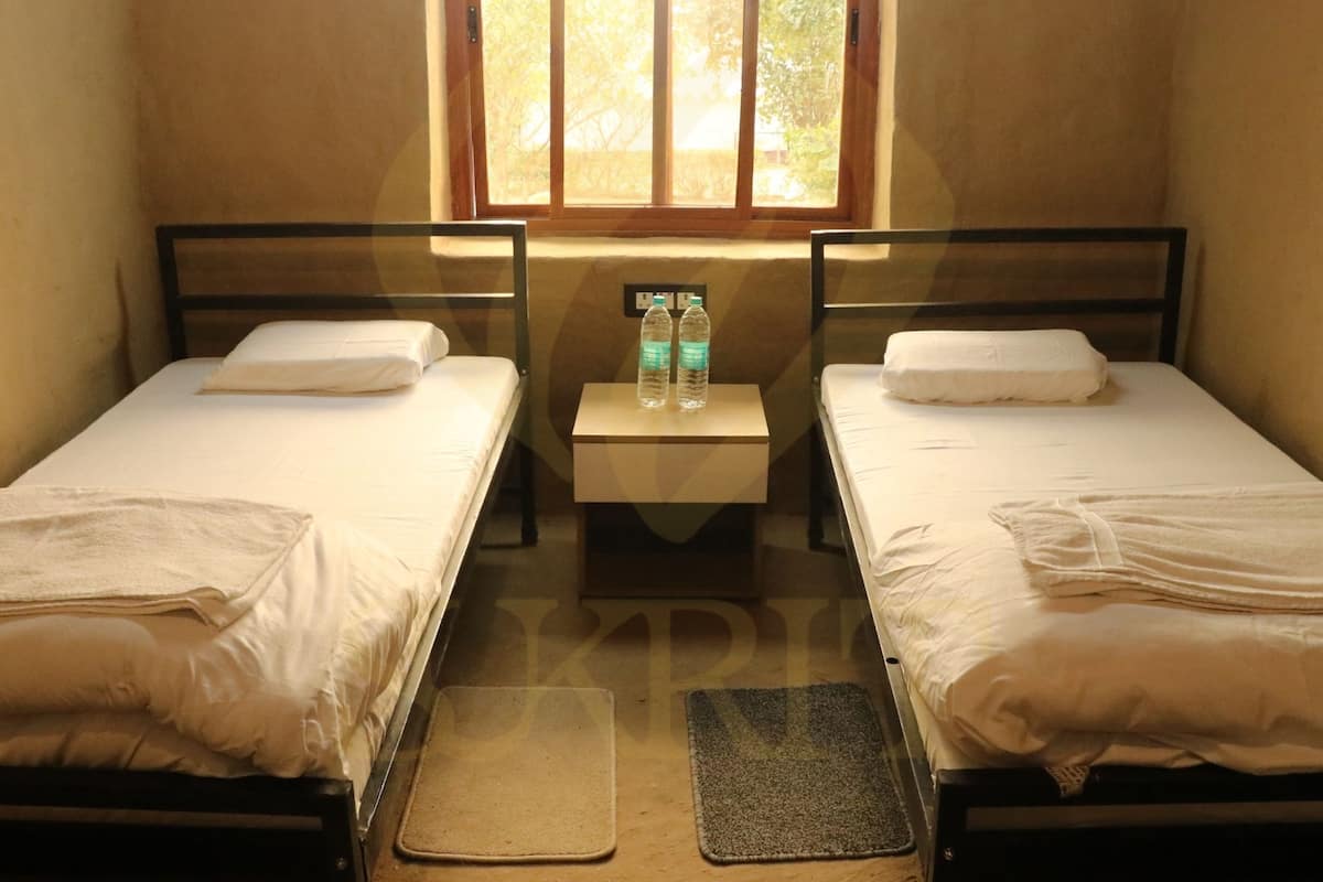 A small, dimly lit room with two single beds, a nightstand with water bottles, and a window with blinds.