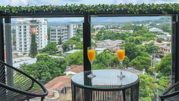 Un balcone con due bicchieri di succo d'arancia su un tavolino di vetro, con vista su un paesaggio urbano ricco di edifici e spazi verdi.
