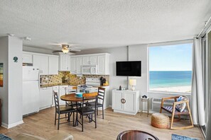 Una cocina con armarios blancos, una mesa de comedor con sillas, un televisor montado en la pared y vistas al océano a través de un amplio ventanal.